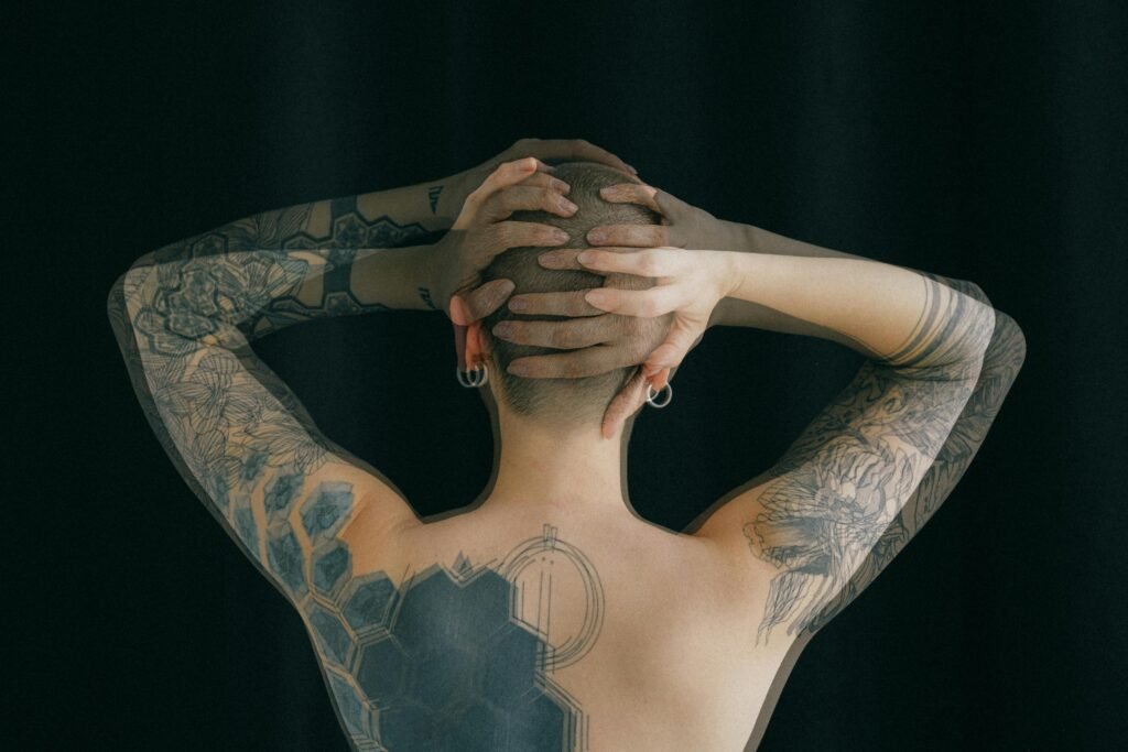 pexels-photo-3851328-3851328 Back view of faceless topless female with tattoos on arm and back standing against black background and holding hands behind head in long expose